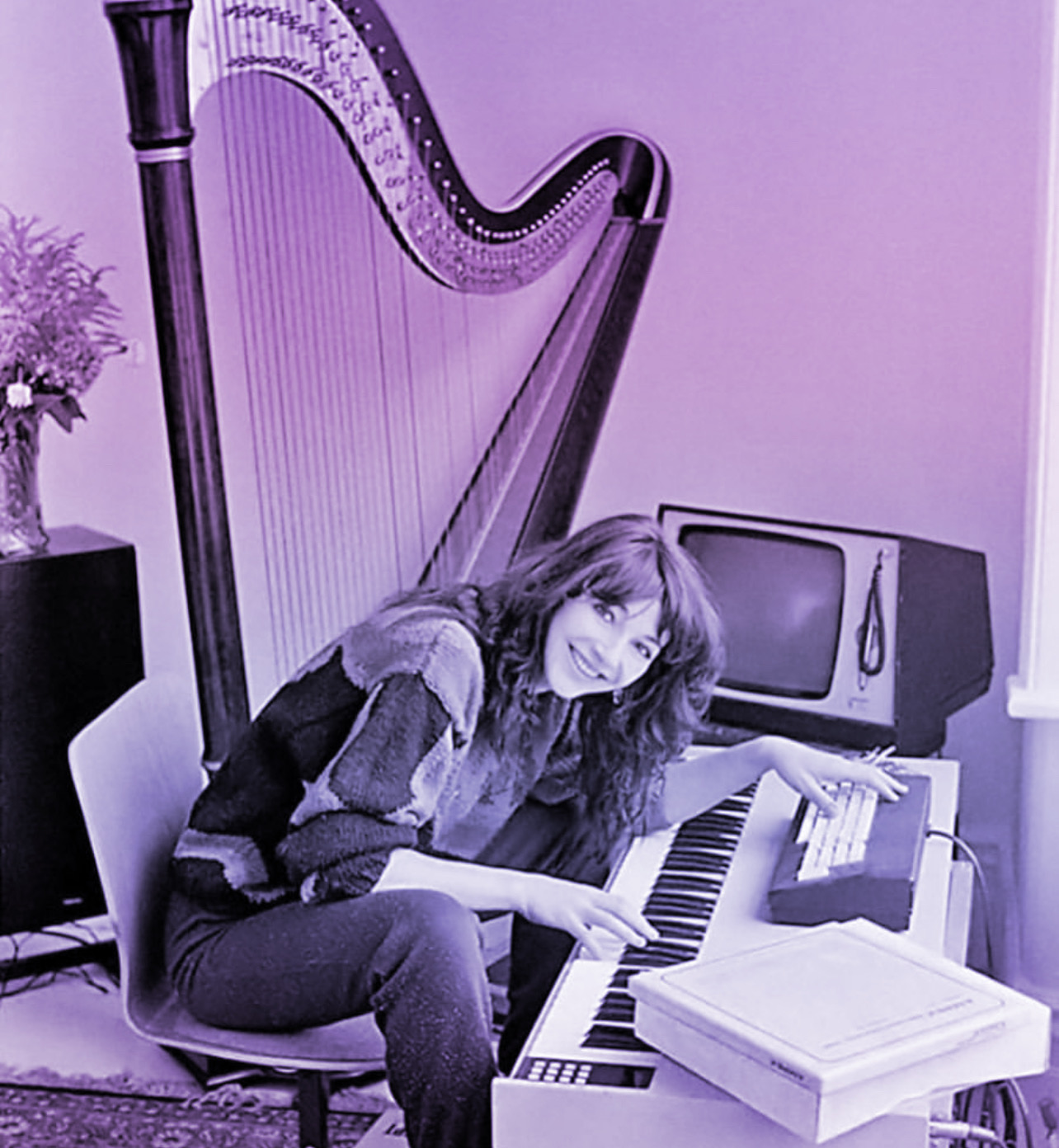 Kate Bush sitting at her Fairlight CMI Synthesizer in her home recording studio with a large harp in the backgroud